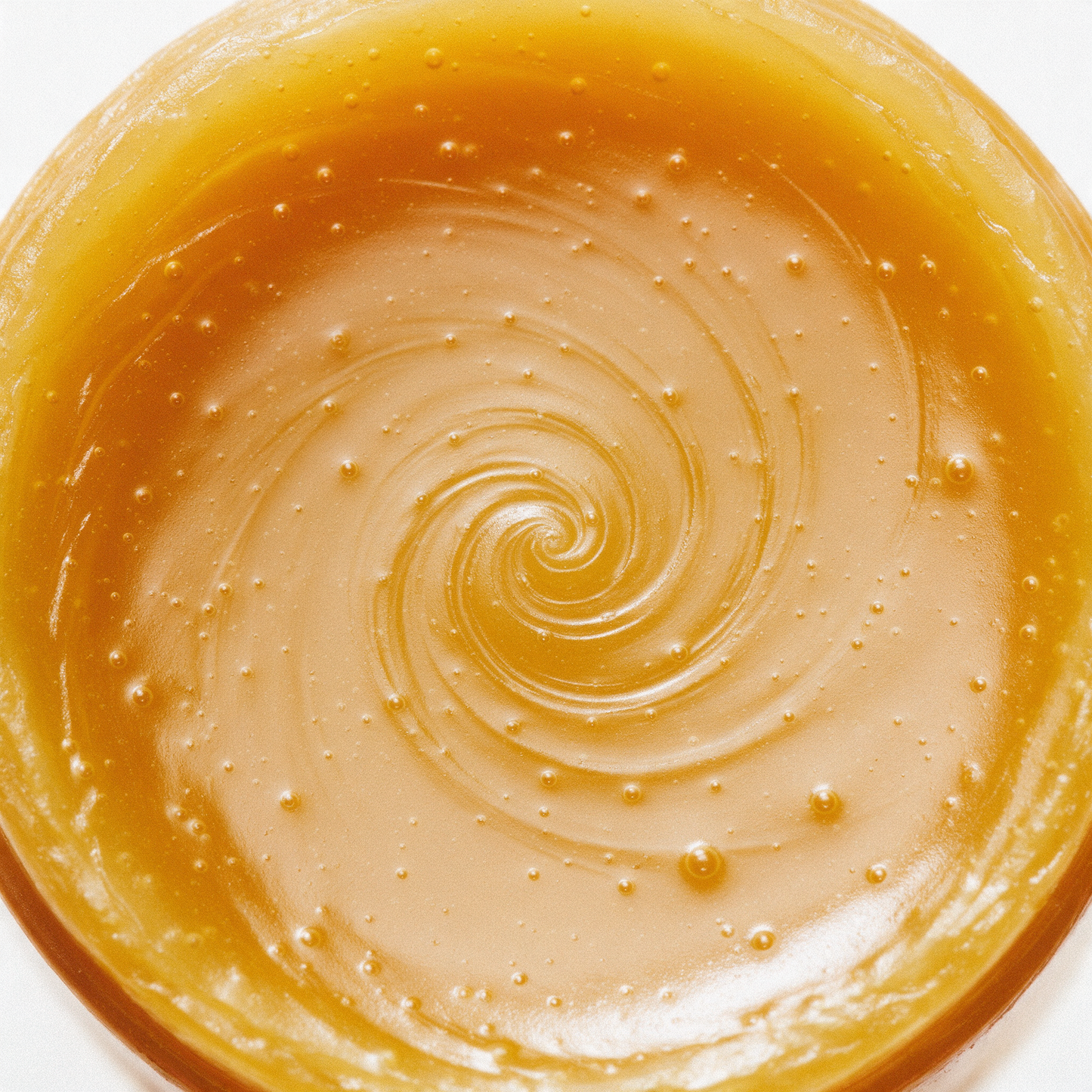 Macro view straight down onto the set surface of a honey-gold beeswax lip balm, showing the pour swirl and tiny suspended bubbles