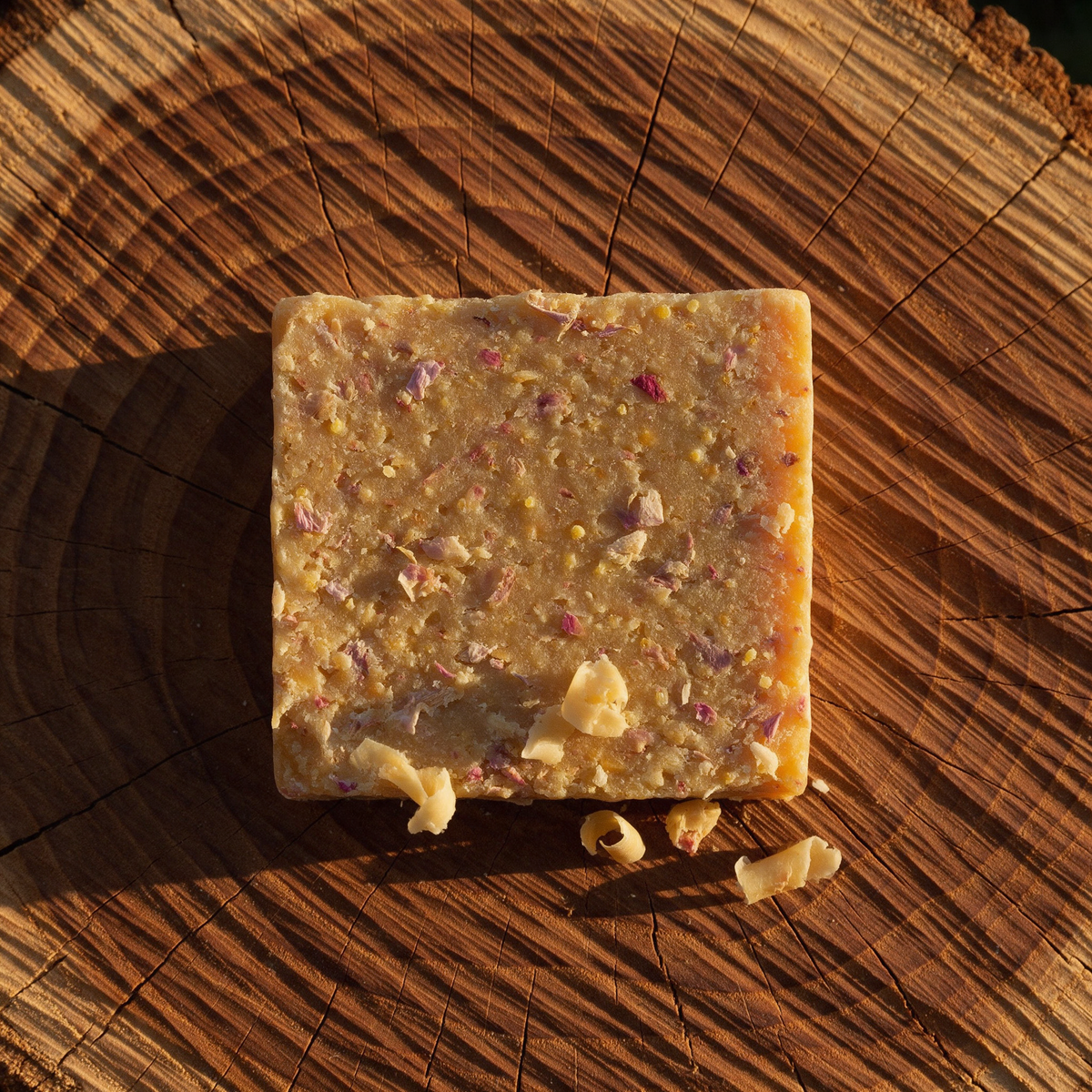 Close-up of a wire-cut soap bar face on sawn teak end-grain, the beeswax grain and its inclusions lit by raking side light, with curled soap shavings beside it