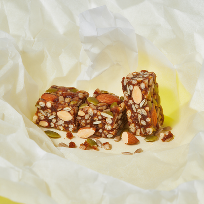 A kids protein energy bar cut into three chunks on crumpled lining paper, the cut faces showing whole almonds, pumpkin and sunflower seeds and soy crispies in a dense date-brown matrix