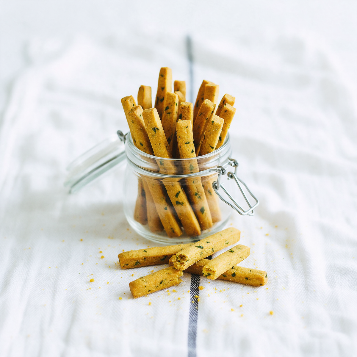 Methi sticks standing upright as a tight bundle in a clip-lid glass jar on white cotton, with five laid crosswise in front and one snapped to show the layered crumb