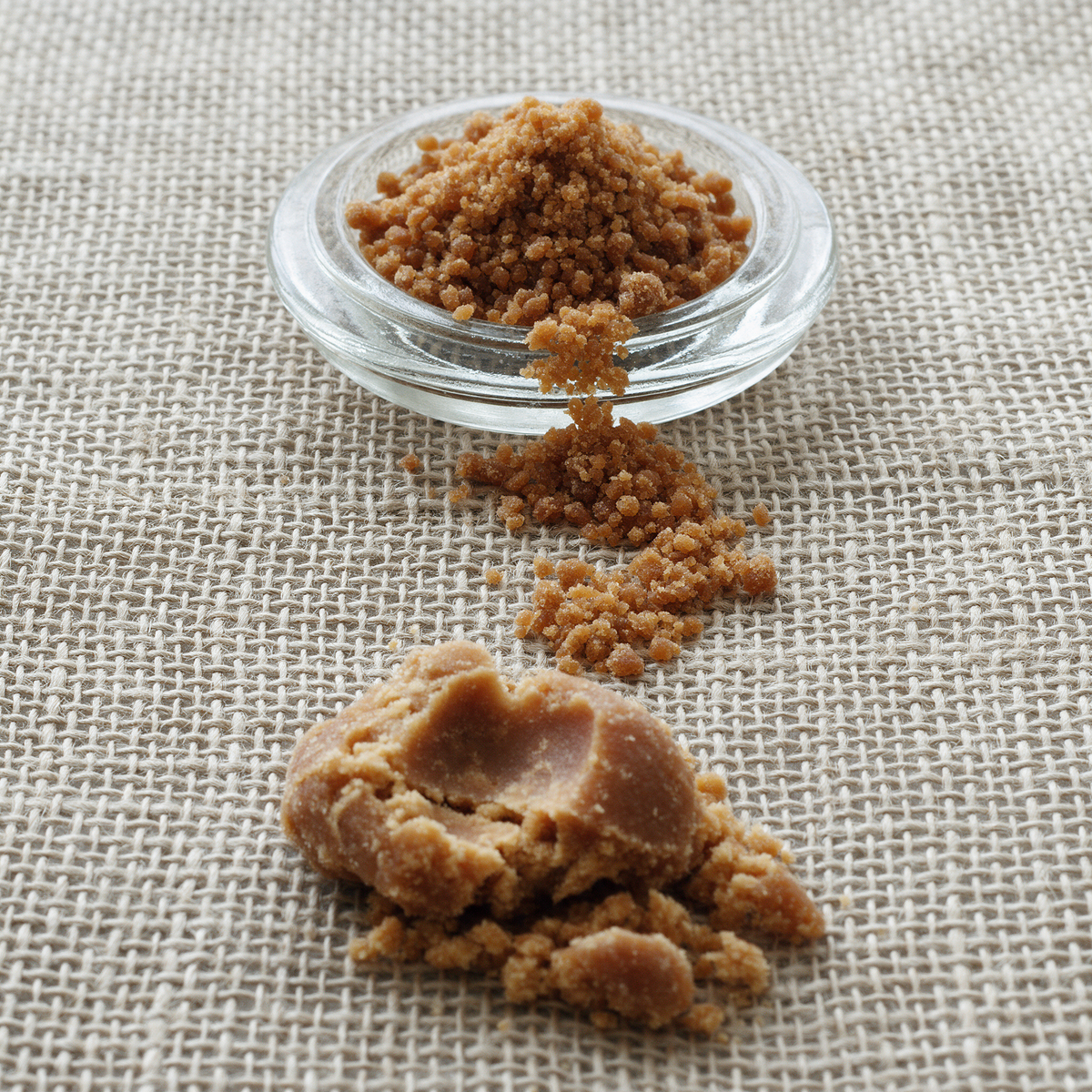 Coarse amber-brown granular jaggery heaped in a pressed-glass dish and trailing across a flax linen runner, with one clump broken open