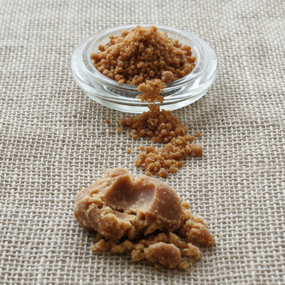 Coarse amber-brown granular jaggery heaped in a pressed-glass dish and trailing across a flax linen runner, with one clump broken open