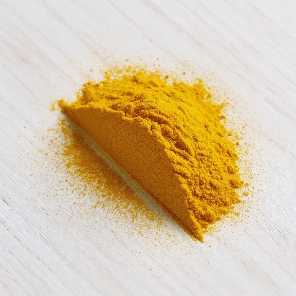 A heap of turmeric powder on a lime-washed timber board struck flat along one edge into a sharp ridge, with a fine dusting halo thrown across the wood