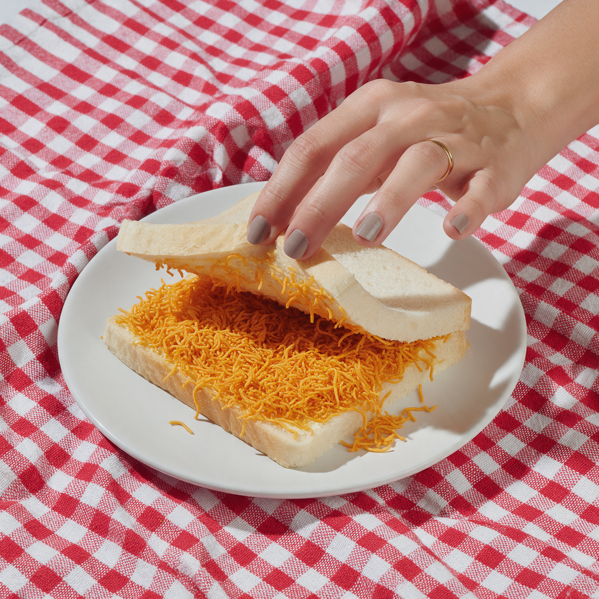 A hand lifting the top slice of a bhujia sandwich on a white plate to show a thick layer of fine orange threads over butter, on a red checked napkin