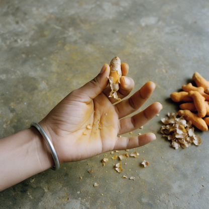A hand rubbing the papery skin off a dried turmeric finger on a cement worktop, turmeric dust in the palm creases, with piles of skinned rhizomes and skins beside