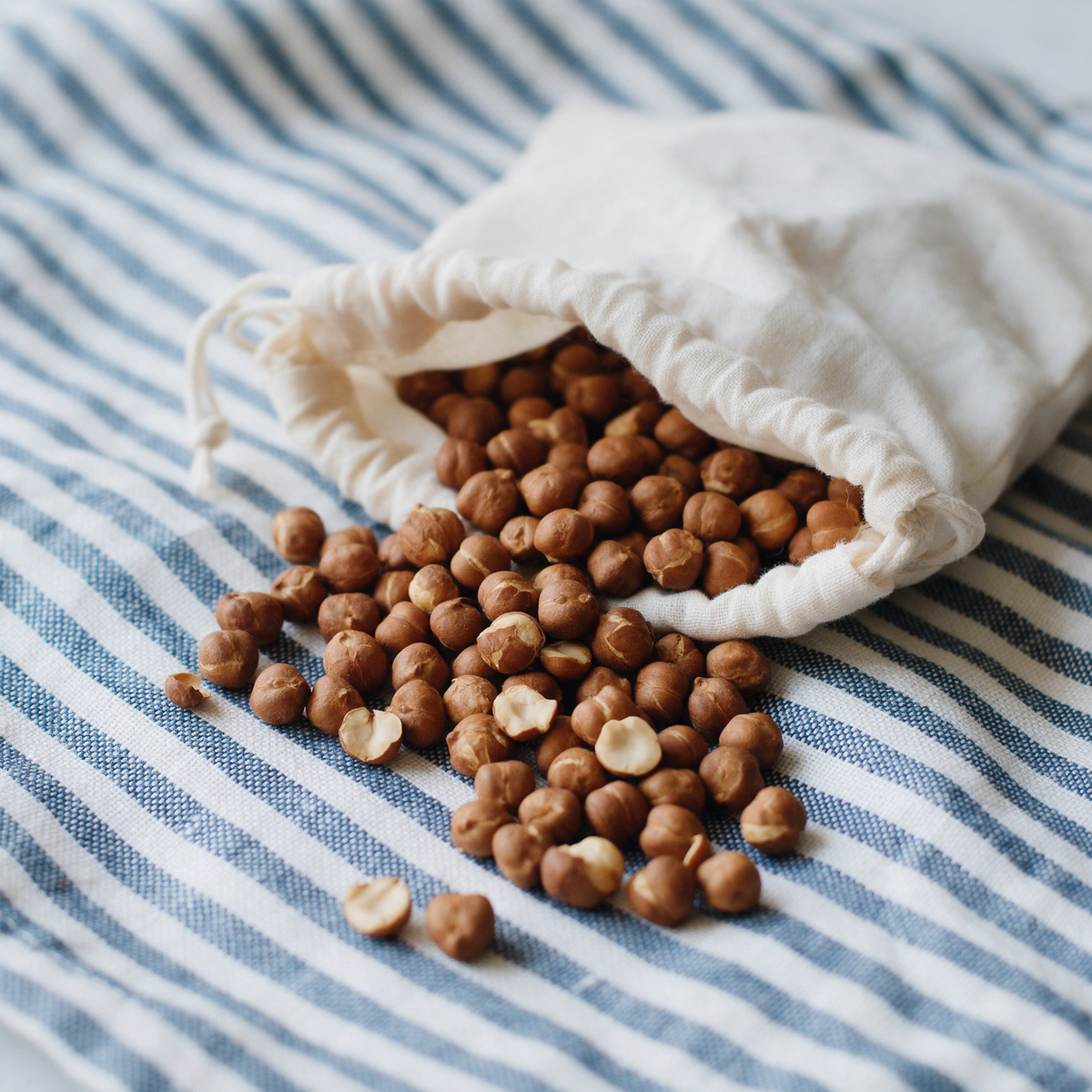 Roasted channa spilling from an open cotton drawstring pouch across striped blue-and-white linen, the skins papery and split and the surface matte and dry