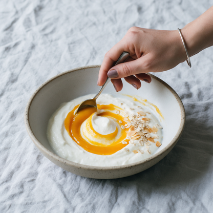 A hand drawing a spiral of Alphonso mango spread through thick set curd in a speckled stoneware bowl, with rolled oats and toasted almond flakes to one side