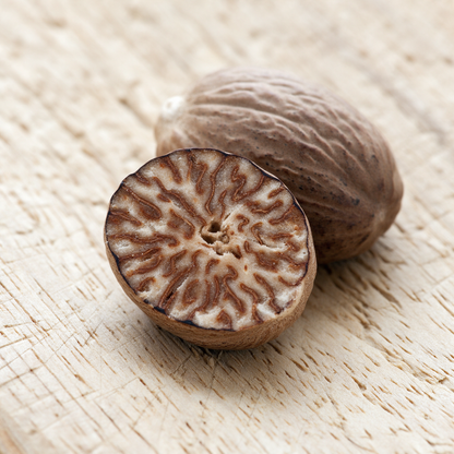 Macro of a nutmeg halved cleanly across with its cut face up on a raw wooden board, the marbled brown-and-cream interior veining beside its whole uncut twin