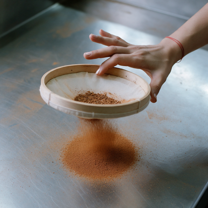 A hand holding a muslin ring sieve above a brushed steel worktop and tapping its rim, fine cinnamon powder falling through into an even circle below