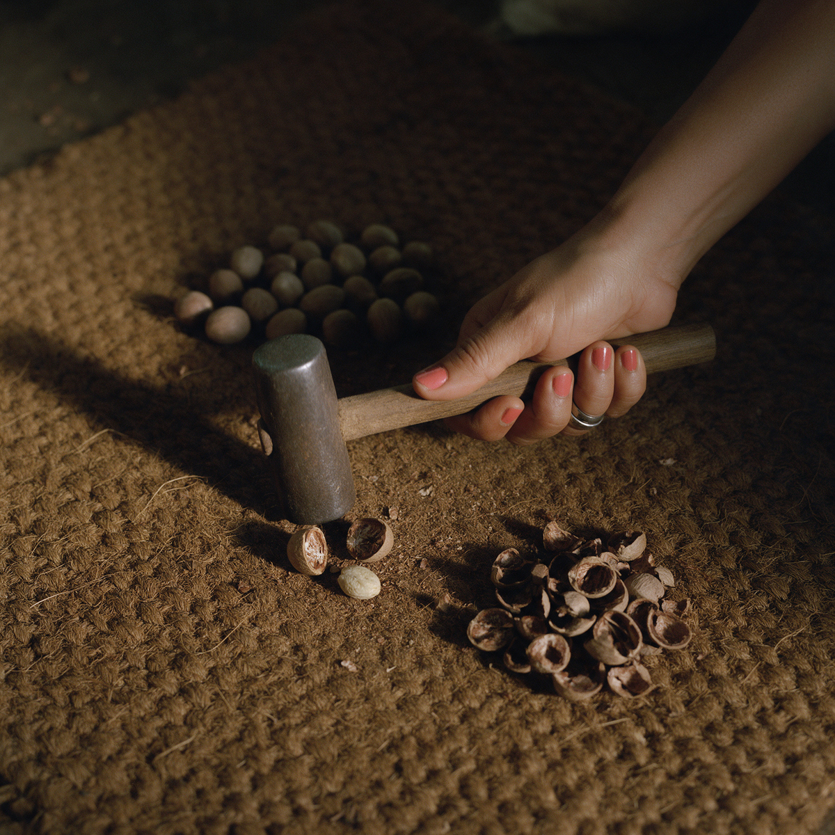 A hand resting a wooden mallet on a coir mat beside a cracked nutmeg shell in two pieces and the freed kernel, with heaps of unshelled nuts and empty shells