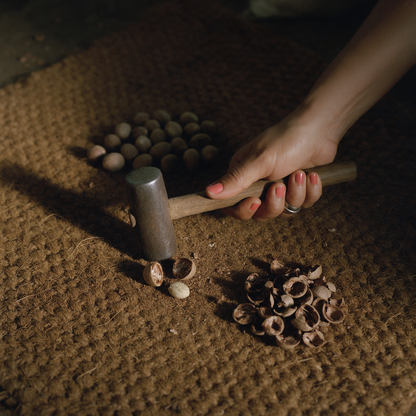 A hand resting a wooden mallet on a coir mat beside a cracked nutmeg shell in two pieces and the freed kernel, with heaps of unshelled nuts and empty shells