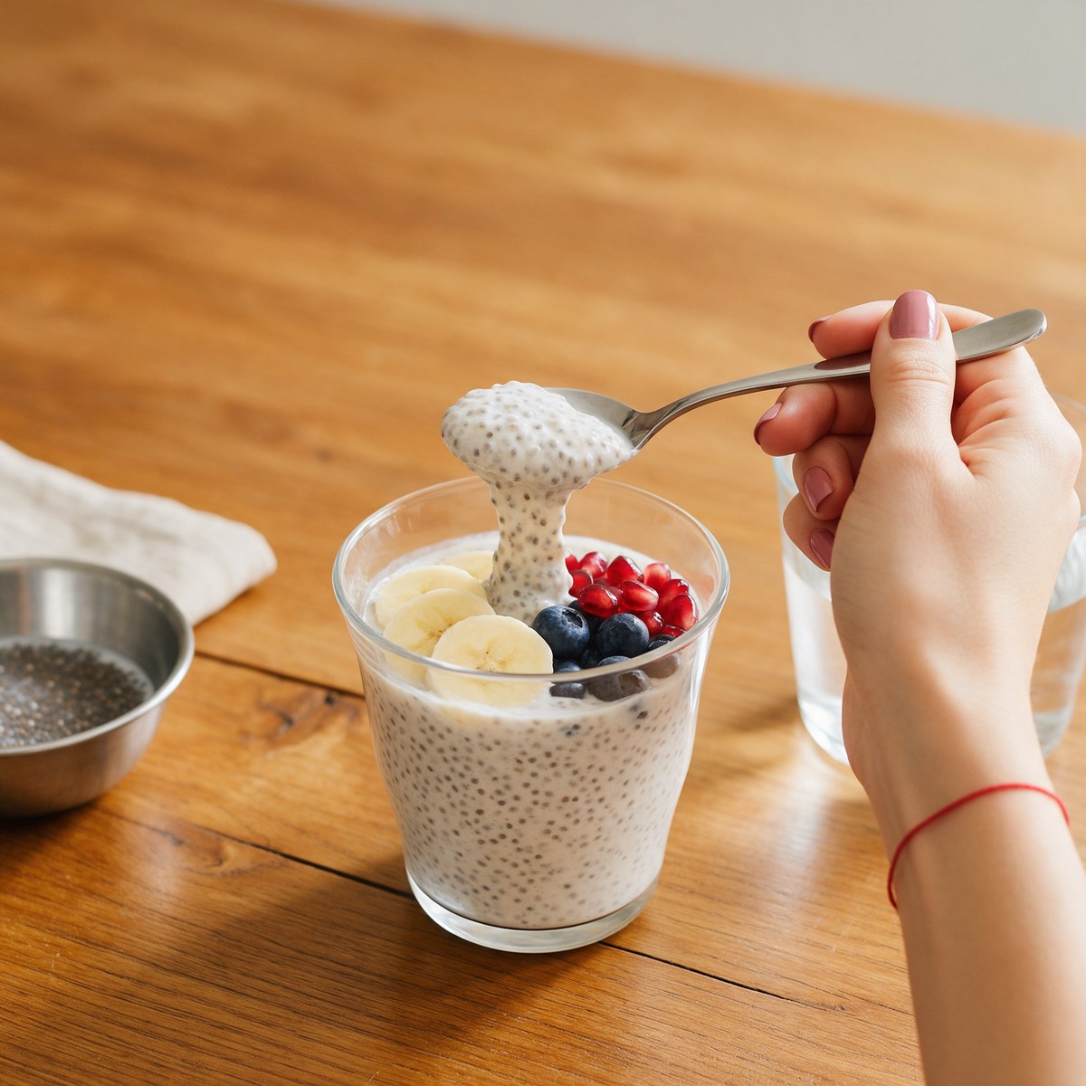 A hand lifting a spoonful of chia pudding topped with banana, blueberries and pomegranate from a glass on a wooden breakfast table