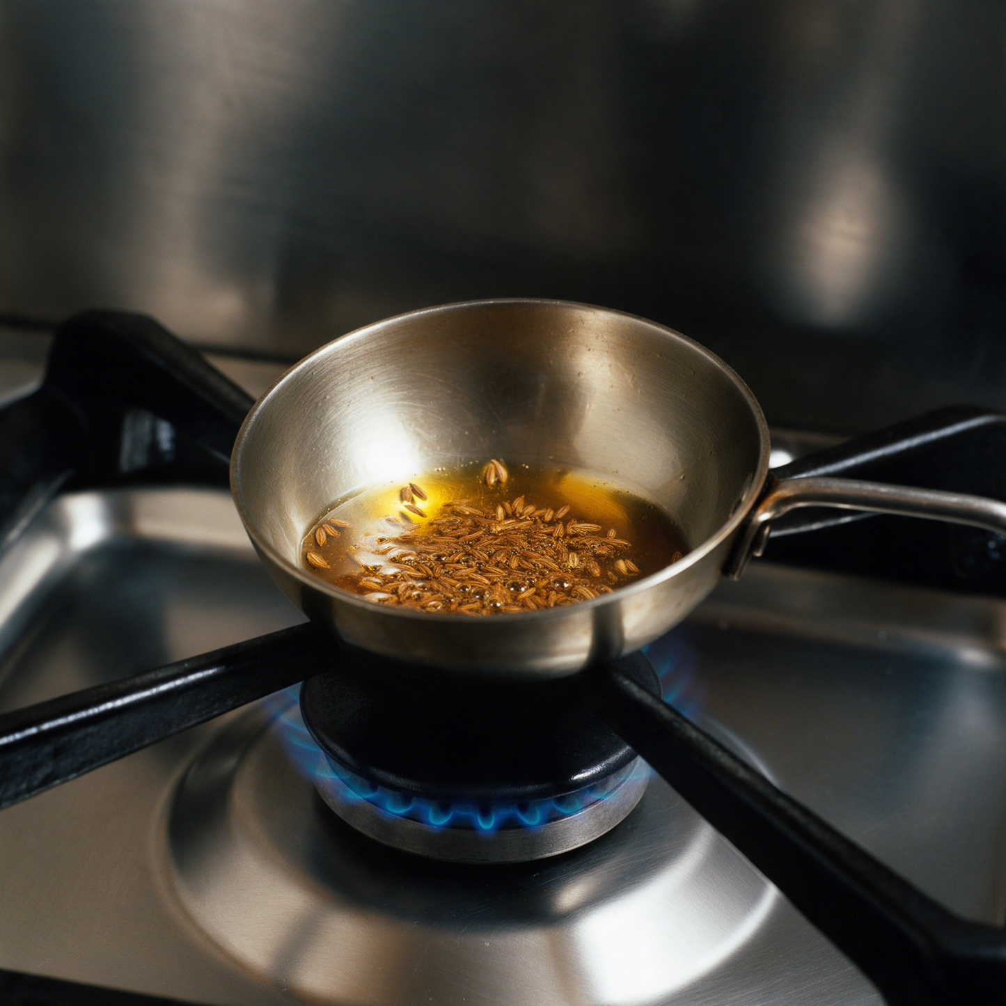 Whole cumin seeds frying in hot ghee in a small steel tempering pan on a gas burner