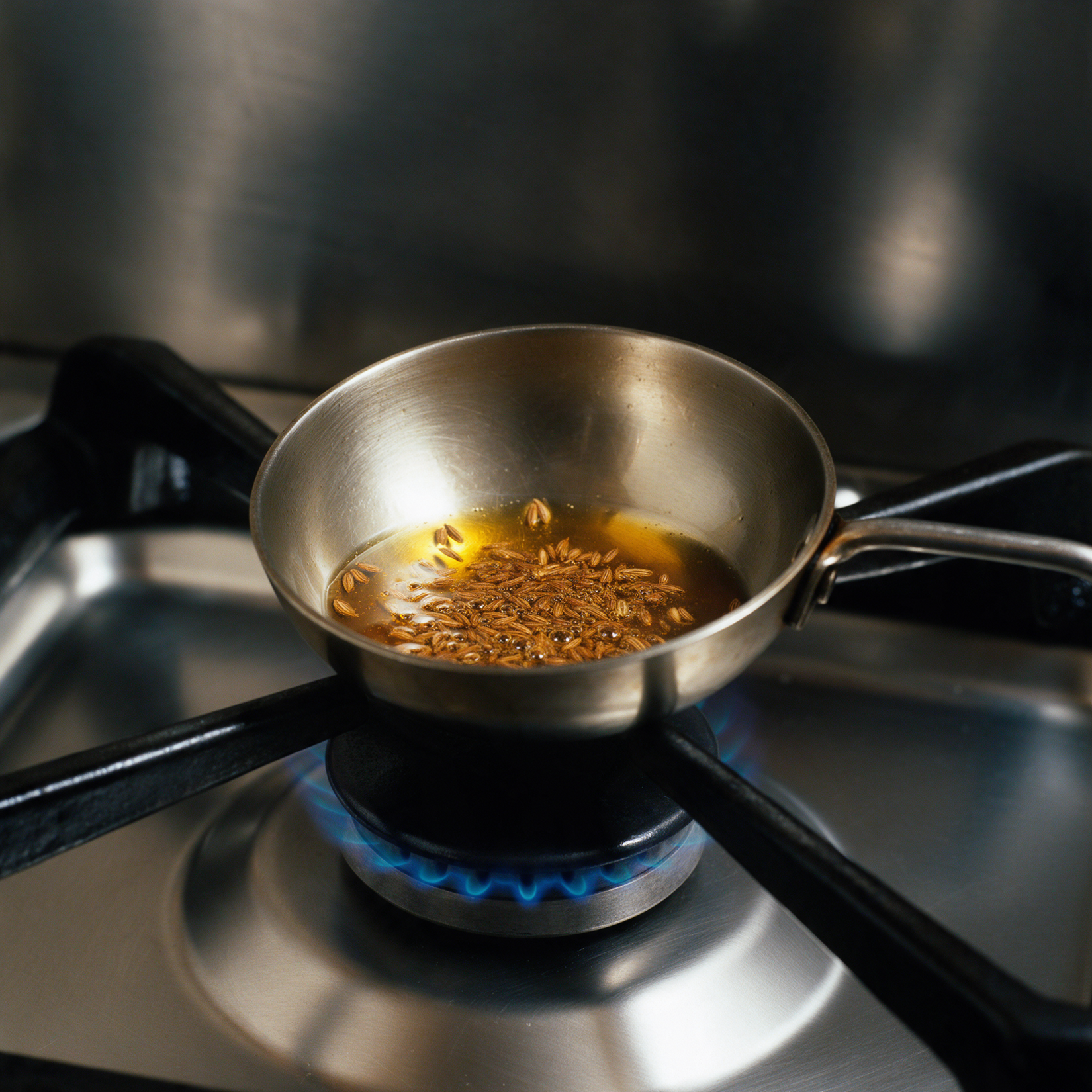 Whole cumin seeds frying in hot ghee in a small steel tempering pan on a gas burner