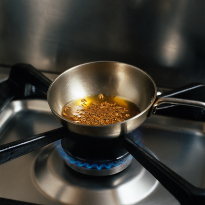 Whole cumin seeds frying in hot ghee in a small steel tempering pan on a gas burner