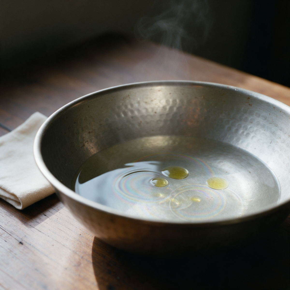 Three drops of lemongrass oil spreading on hot water in a hammered steel bowl for steam inhalation, a folded cotton cloth beside it