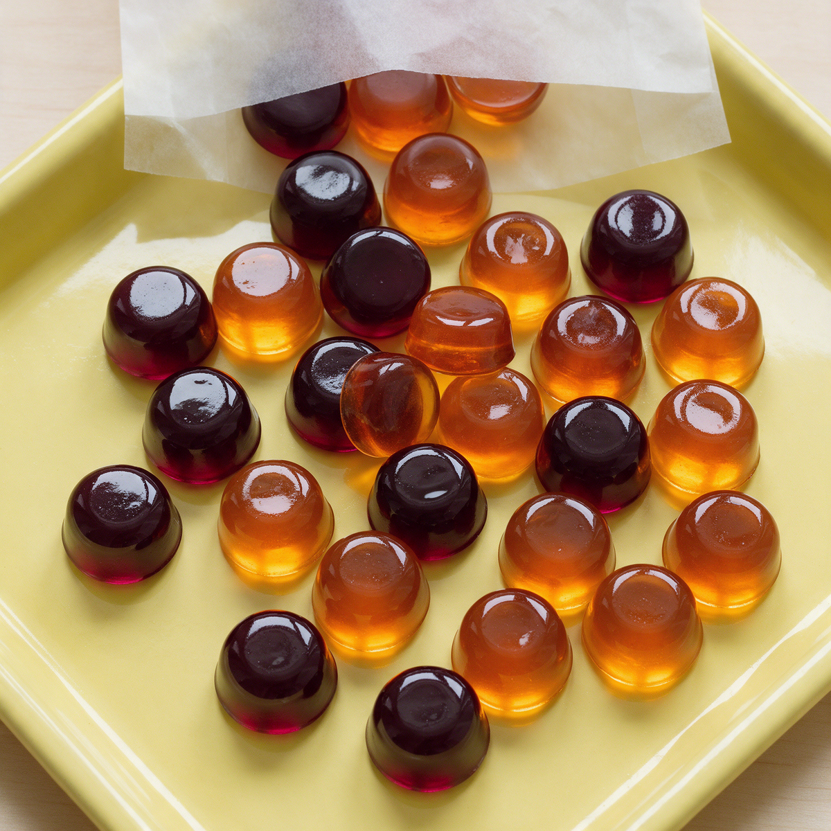 Round translucent maroon and amber fruit gummies spilling from a plain paper sleeve across a lemon-yellow enamel tray