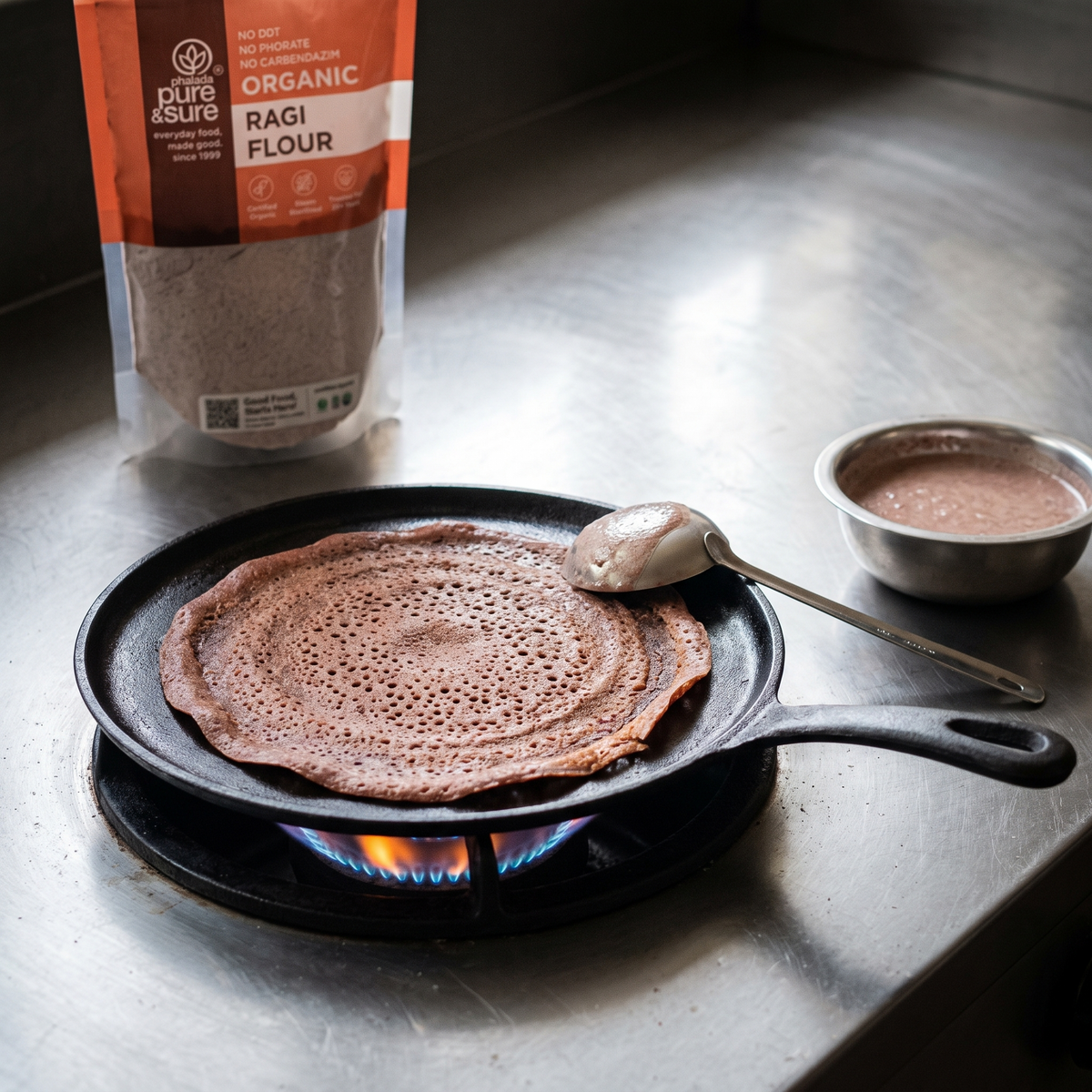 A ragi dosa spread on a hot cast-iron tawa with the ragi flour pack behind