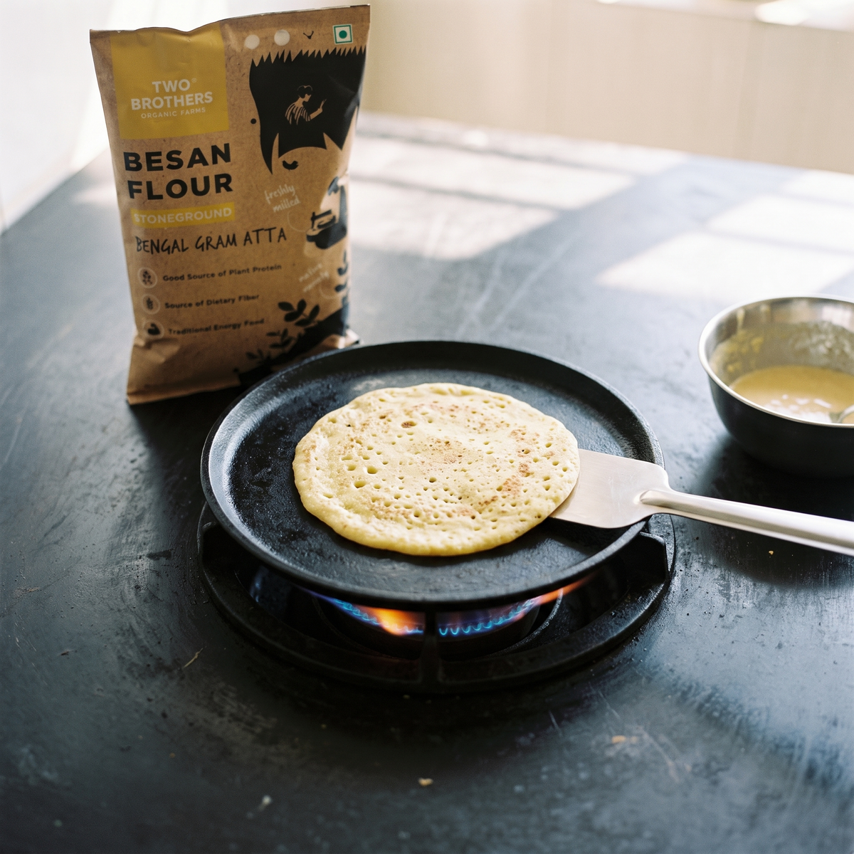 A besan chilla cooking on a cast-iron tawa with the stoneground besan flour pack behind
