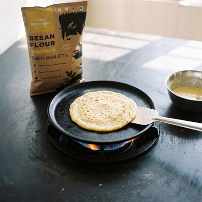 A besan chilla cooking on a cast-iron tawa with the stoneground besan flour pack behind