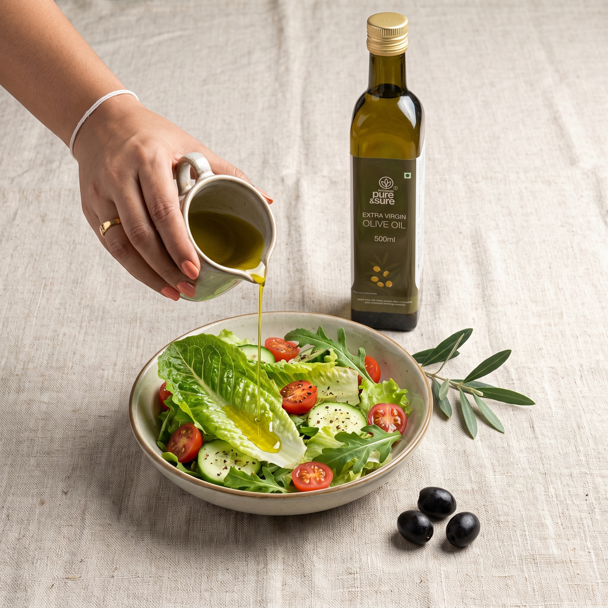 Extra virgin olive oil drizzled from a ceramic pourer over a bowl of salad leaves, with the Pure & Sure 500ml bottle standing beside it