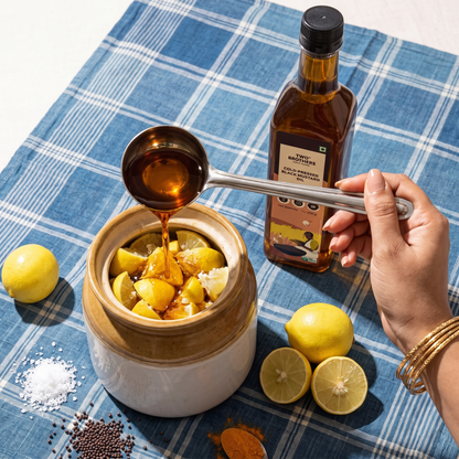 Cold-pressed black mustard oil being poured into a ceramic jar of lemon wedges while making nimbu achar, with salt, mustard seeds and turmeric on an indigo check cloth