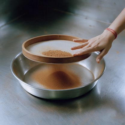 A wooden ring sieve resting on the rim of a steel parat, a hand steadying it while fine cinnamon powder falls through into a cone in the tray below