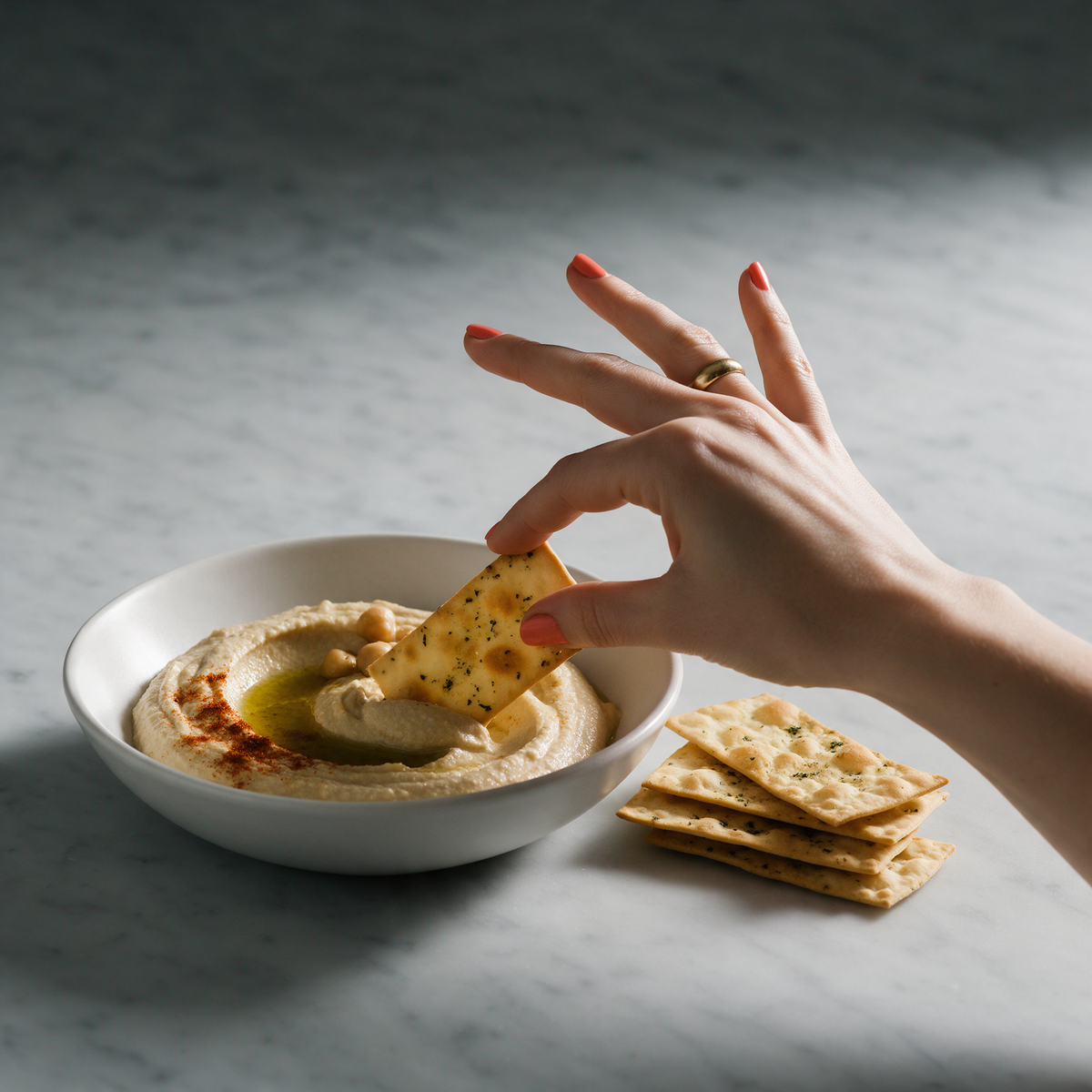 A hand dipping a HridiKa's gluten free herbed aglio lavash cracker into a bowl of hummus dressed with olive oil and paprika
