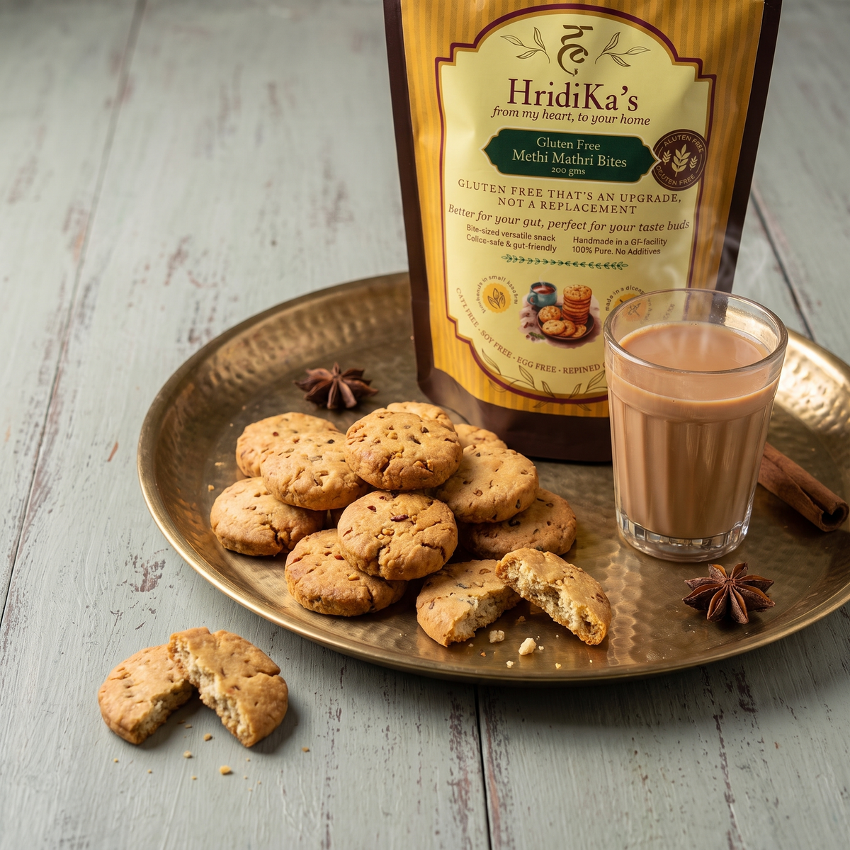 HridiKa's gluten free methi mathri bites piled on a hammered brass plate with a glass of chai, star anise and cinnamon, the pack standing behind